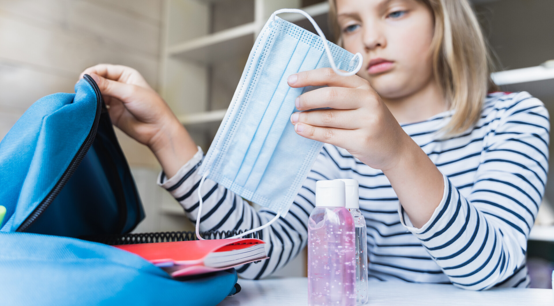 Little girl packing a backpack with a bottle of sanitizer and a face mask