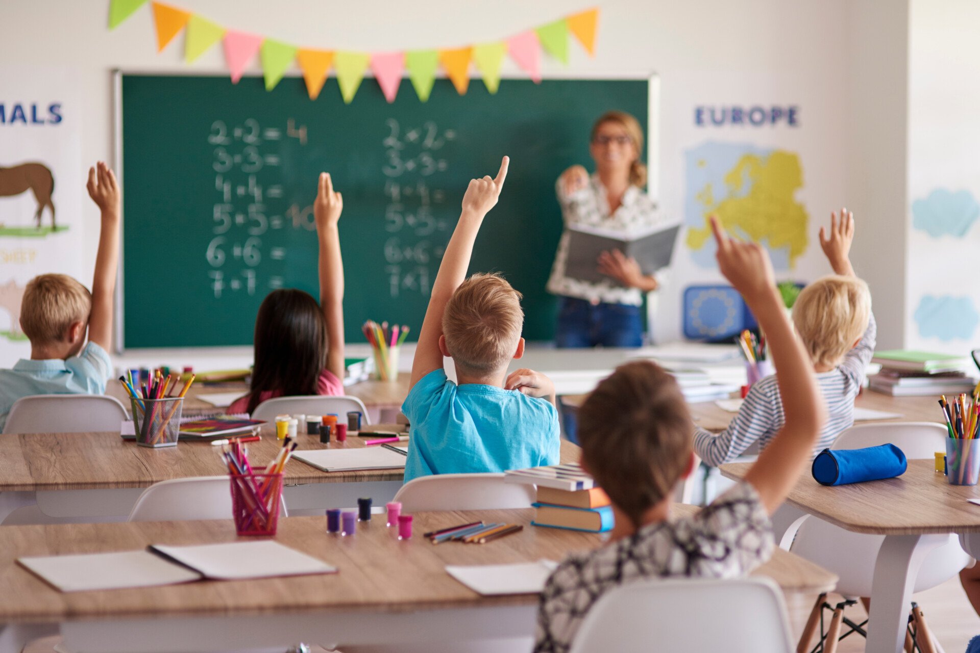 Classroom full of children with their hands raised. Teacher at the front chooses a child to answer the question.