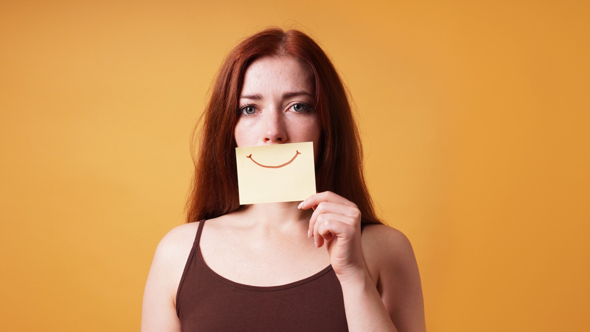 young woman with sad eyes hiding real emotion by covering mouth with fake smile drawn on paper