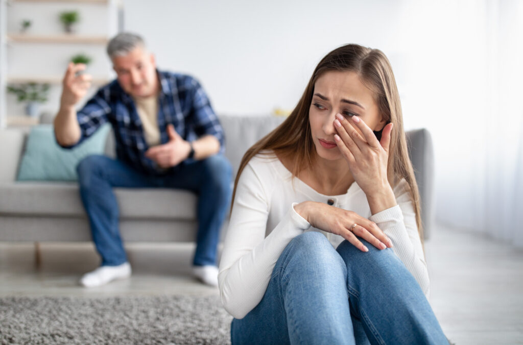 A woman sat on the floor crying while a man behind her is shouting at her