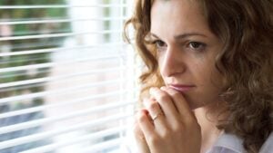 A socially anxious woman looking out of the blinds in her house with concern.