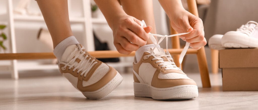 Woman in stylish sneakers tying shoe laces in shop